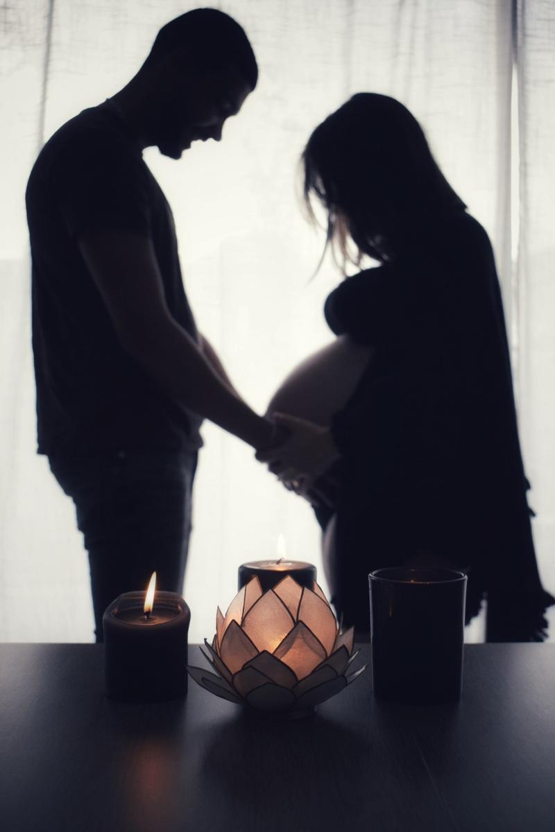 Silhouette d'un couple à contre-jour, les mains sur le ventre, derrière des bougies et un photophore lotus